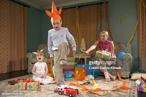 young boy (6-7) about to destroy toy car, girl (3-4) crying, mother sitting in armchair - party inside stockfoto's en -beelden