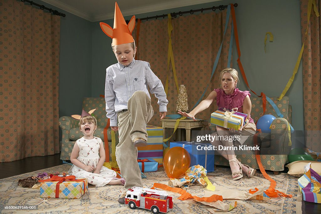 Young boy (6-7) about to destroy toy car, girl (3-4) crying, mother sitting in armchair