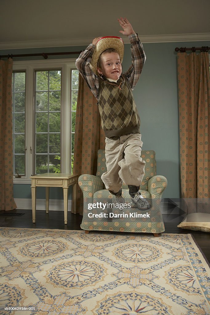 Young boy (6-7 years) wearing cowboy hat and boots, jumping from armchair in living room, portrait