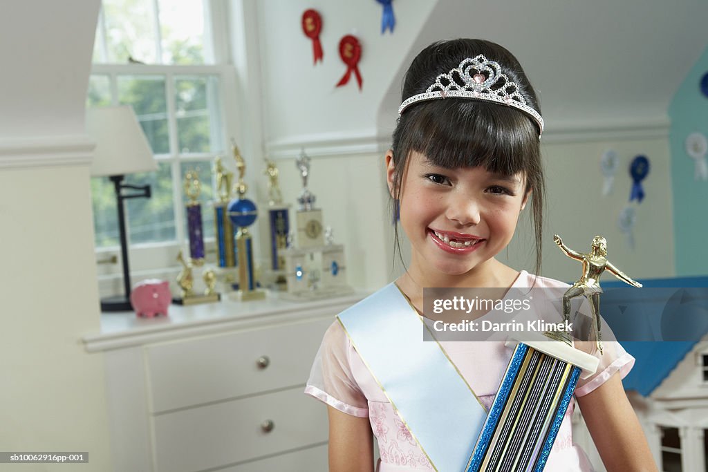 Portrait of young girl (8-9 years) indoors, wearing sash and tiara, holding prize, smiling