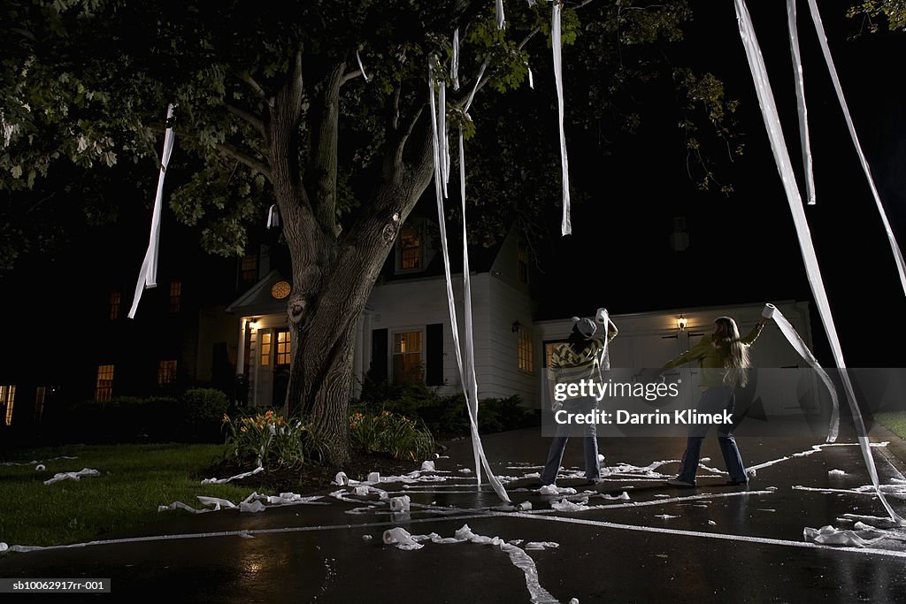 Two girls (10-11, 12-13) throwing toilet paper into tree in front of house, night