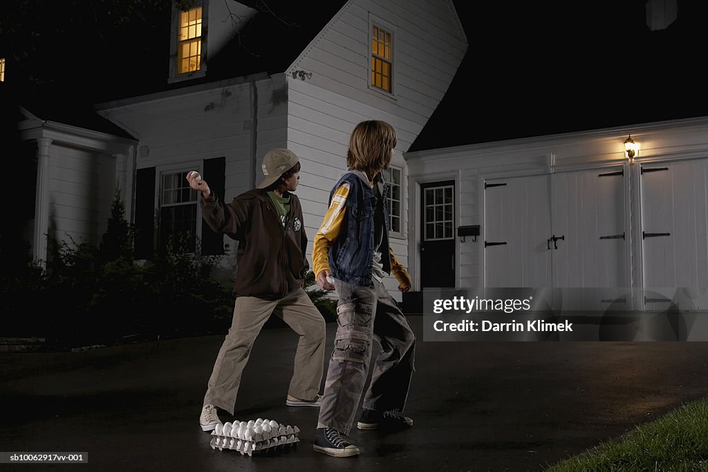 Two boys (12-13) standing in driveway, throwing eggs at house, night, rear view