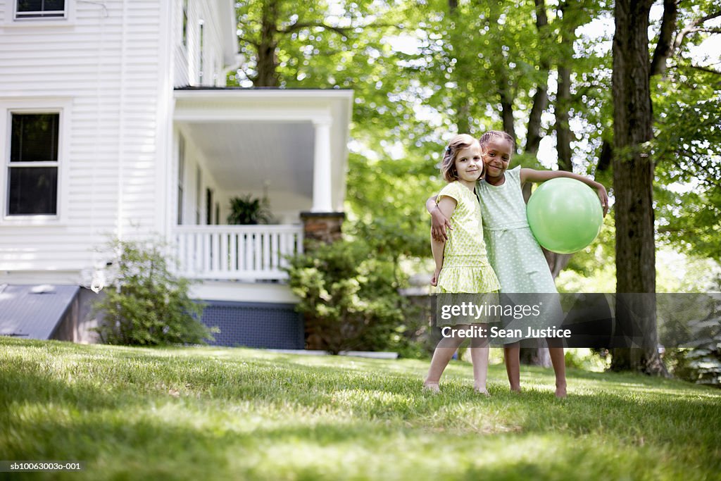 Two young girls (7-8 years) standing together on lawn, one holding green ball, portrait
