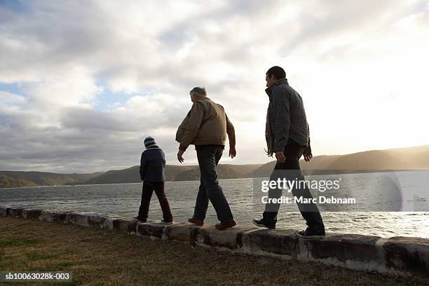 boy (10-11), grandfather and father walking on wall at edge of lake, rear view - father and son walking stock pictures, royalty-free photos & images