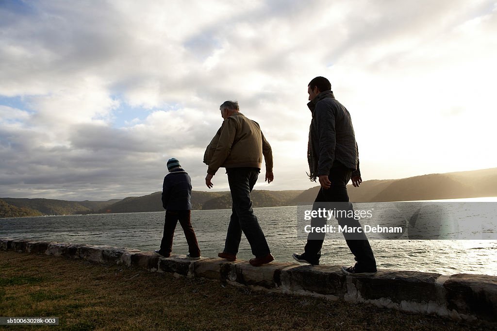 Boy (10-11), grandfather and father walking on wall at edge of lake, rear view