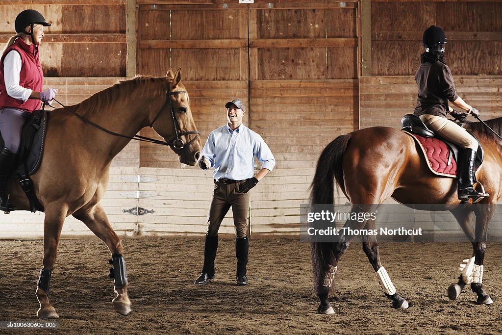 Young man standing between to riders and their horses in a training stable, smiling