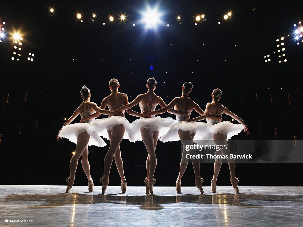 Five ballerinas en pointe on stage, arms around each other, rear view