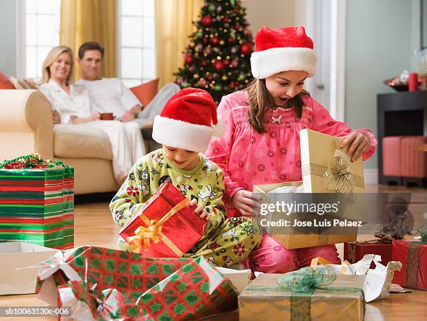 boy (6-7 years) and girl (8-9 years) unwrapping christmas presents in living room, parent sitting in background - open day 7 stockfoto's en -beelden