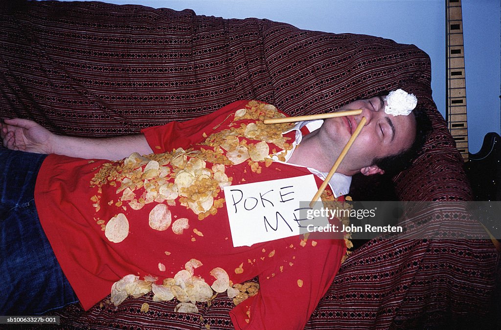 Young man asleep on sofa covered in crisps wearing sign