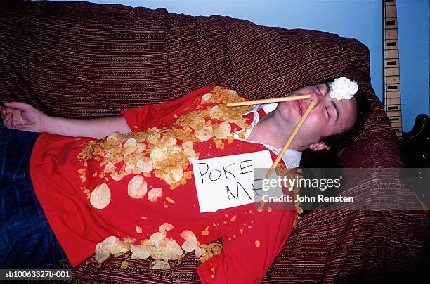 young man asleep on sofa covered in crisps wearing sign - schaamte stockfoto's en -beelden