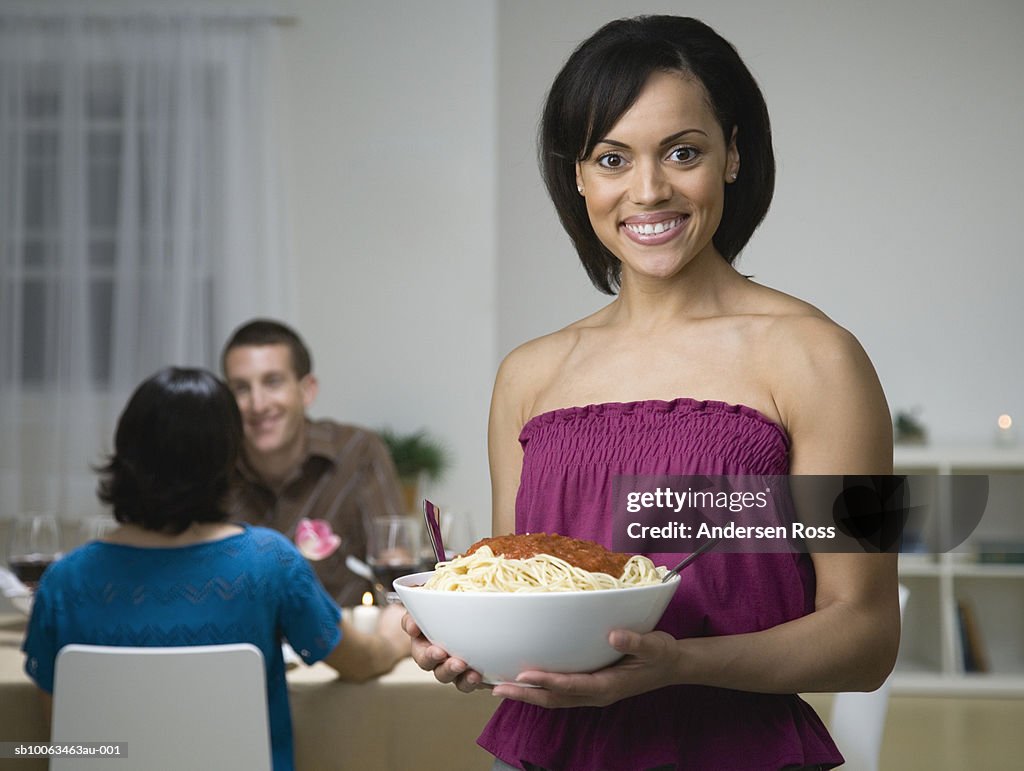 Young woman holding serving bowl of spaghetti, others at dinner table, at home