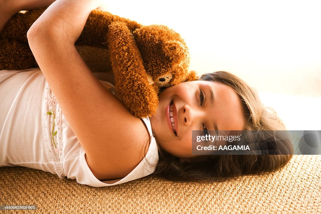 Girl (8-9) lying on floor hugging teddy bear, smiling, portrait