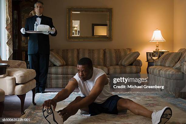 mature man exercising on carpet, butler waiting with water on tray, in living room - gambe divaricate foto e immagini stock