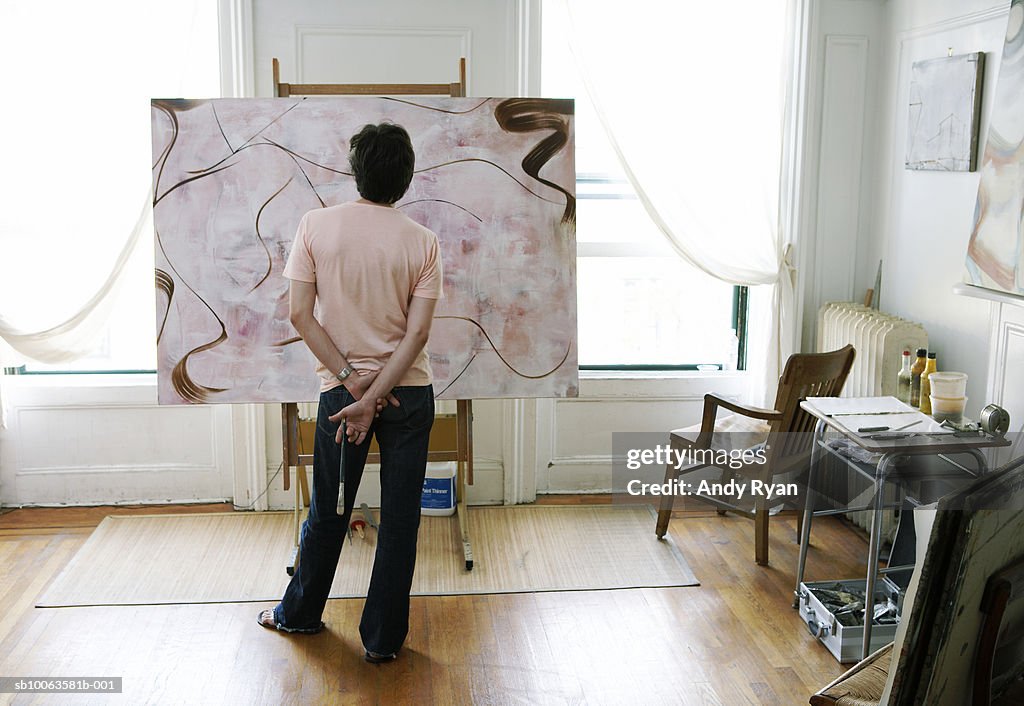 Man holding paint brush looking at painting on easel, rear view
