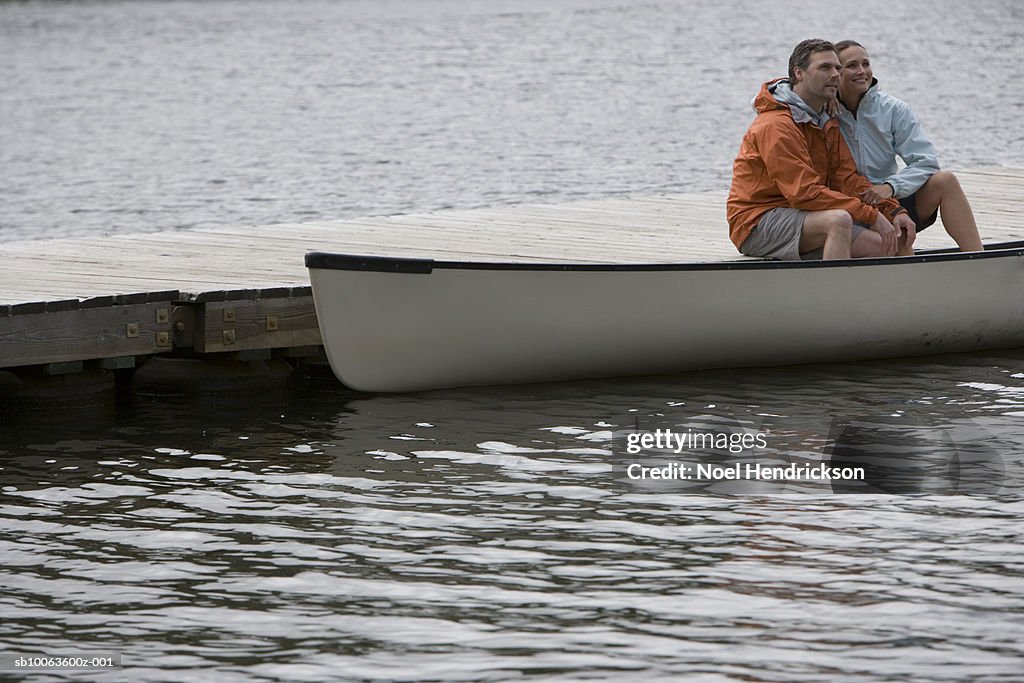 Couple sitting on pier with legs in canoe, looking at view