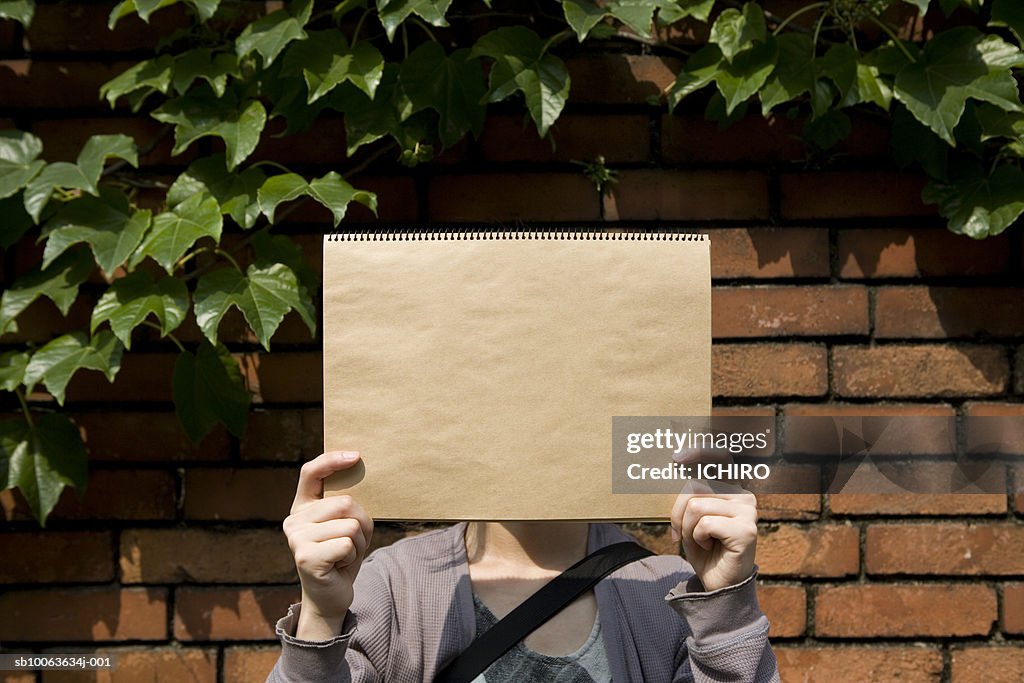 Woman holding sketch pad over face, standing in front of brick wall