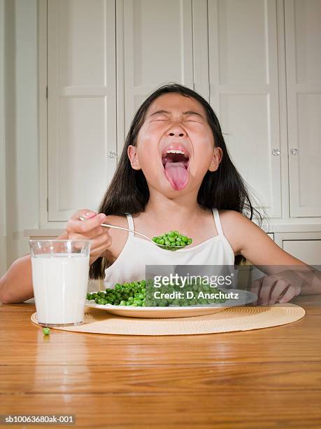 girl (8-9) sitting at table with plate of green peas, sticking out tongue - girl open mouth stockfoto's en -beelden