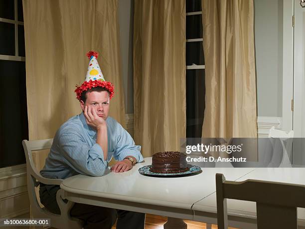 mid adult man in party hat, sitting at table in front of birthday cake - lonely stock-fotos und bilder