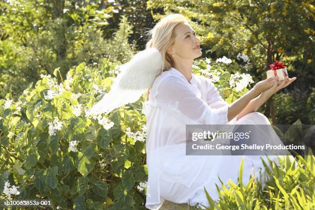 woman wearing angel wings holding small gift, sitting in garden, side view - angel white dress stock pictures, royalty-free photos & images