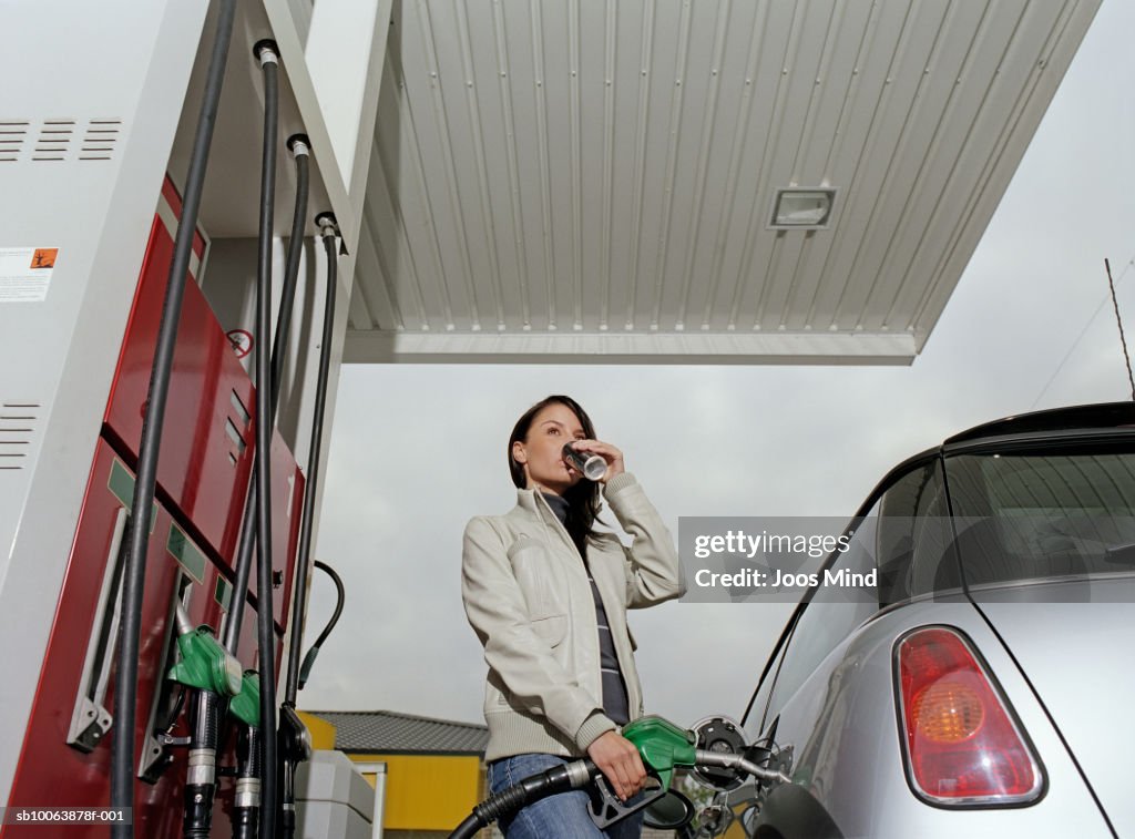 Young woman filling car at petrol station drinking from tin, low angle view
