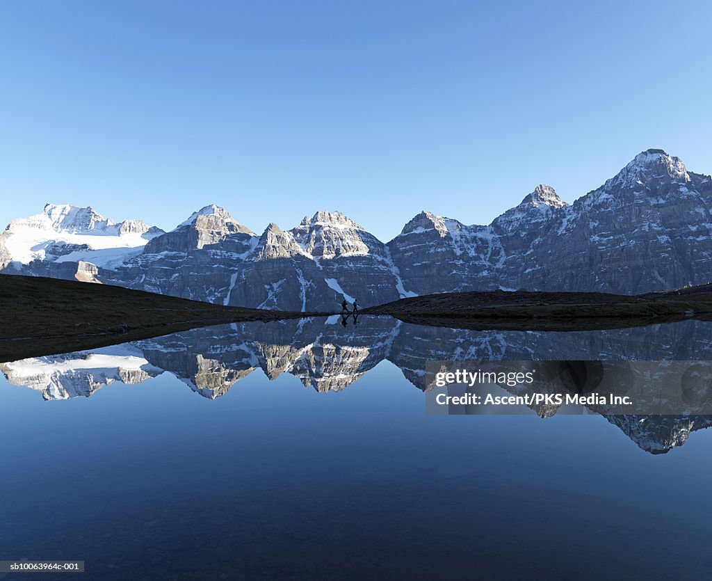 Snowcapped mountains reflecting in lake water, two hikers walking in distance