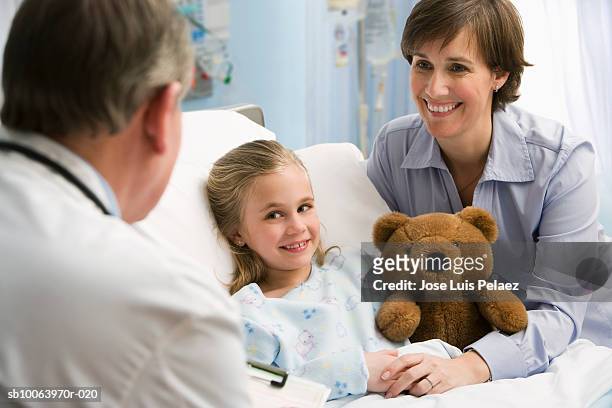 male doctor smiling at mother and girl (6-7) holding teddy bear - adult male hospital bed stock-fotos und bilder