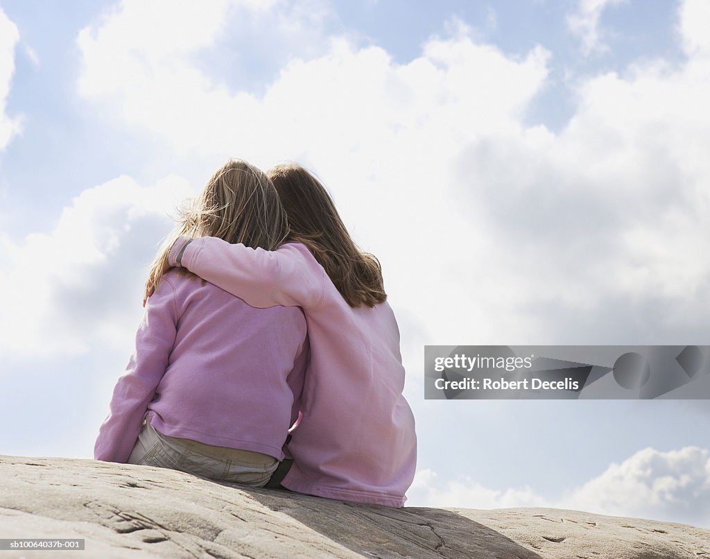 Two girls (6-9) sitting on rock with arms around, rear view