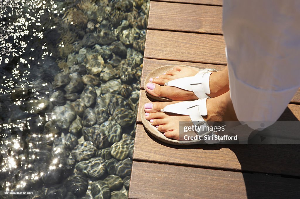 Woman wearing sandals standing on decking by pond, low section, high angle view