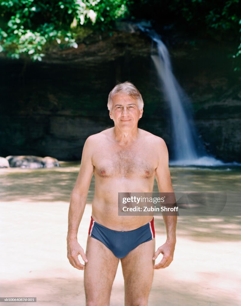 Senior man standing in pond, waterfall in background, smiling, portrait