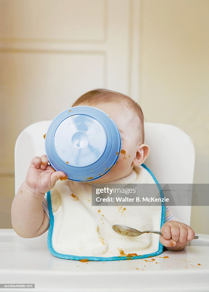 Baby girl (6-7 months) holding bowl to face sitting in high chair