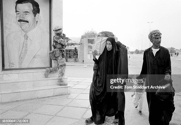 Wary Iraqi family pass a British Royal Marine Commando near a mural of Saddam Hussein on the day Basrah was liberated.
