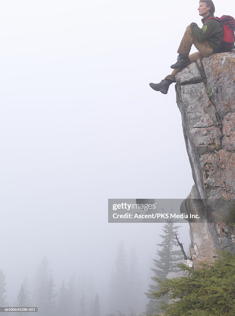 Mountaineer sitting on edge of cliff, fog and trees below