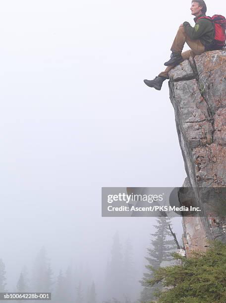 mountaineer sitting on edge of cliff, fog and trees below - cliff side stock pictures, royalty-free photos & images