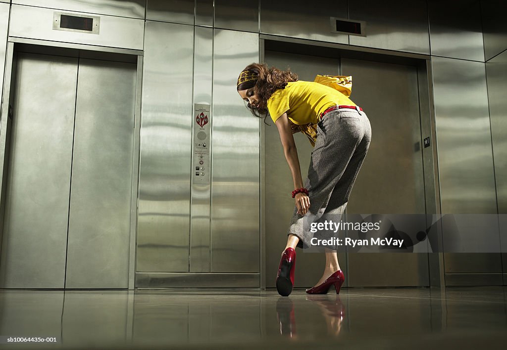 Young woman standing in front of elevators inspecting trousers and shoe, rear view