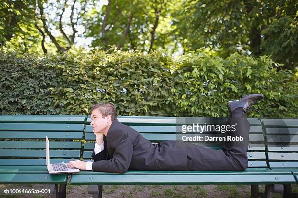 young businessman lying on bench in park, using laptop, side view - china foto e immagini stock