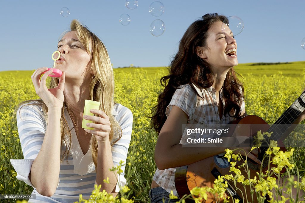 Two young women in field, one playing guitar, one blowing bubbles