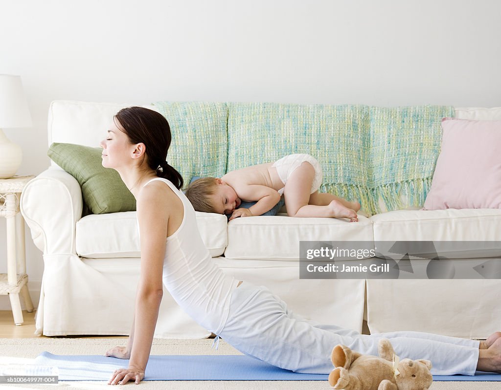 Mother doing yoga, son (21-24 months) on sofa in background