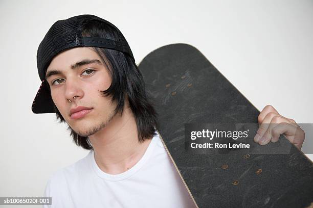 teenage boy (14-15) holding skateboard on shoulder, wearing baseball cap askew, portrait - teenage boy in cap posing stock pictures, royalty-free photos & images