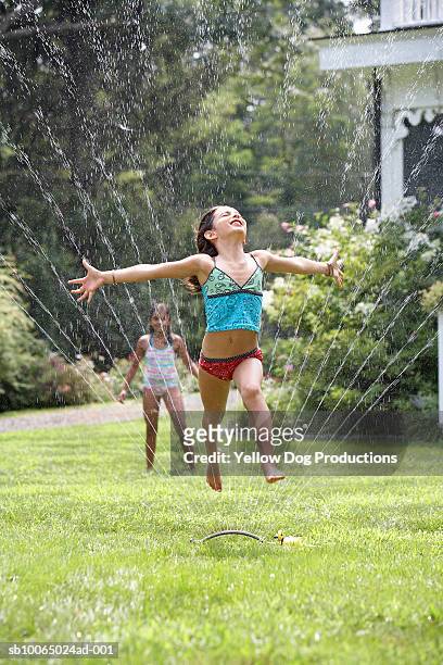 two girls (8-9) playing in garden, one jumping through sprinkler - jumping sprinkler stock pictures, royalty-free photos & images