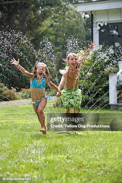 boy and girl (5-6) jumping through sprinkler in garden - jumping sprinkler stock pictures, royalty-free photos & images