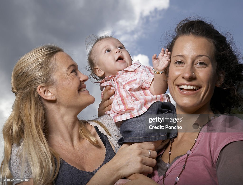 Germany, Bavaria, two women holding baby (3-6 months) outdoors, low angle view