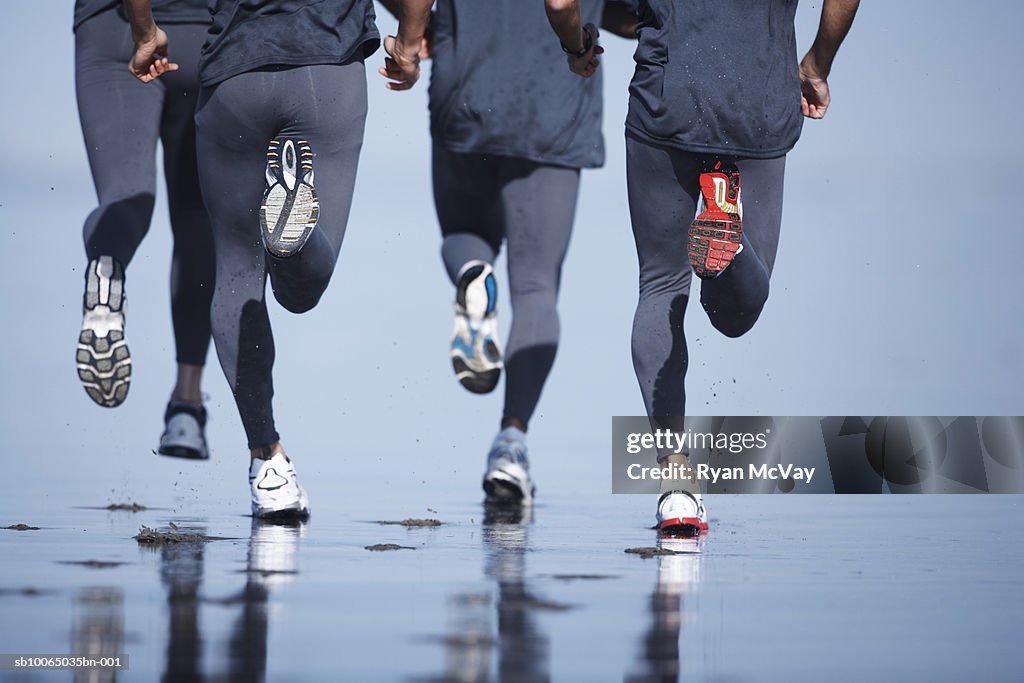 Four men jogging on beach, low section, rear view