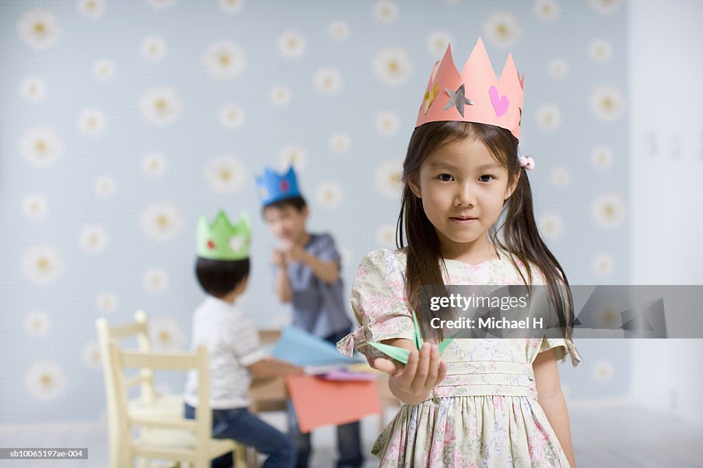Three children (4-5) making origami in room, focus on girl in foreground