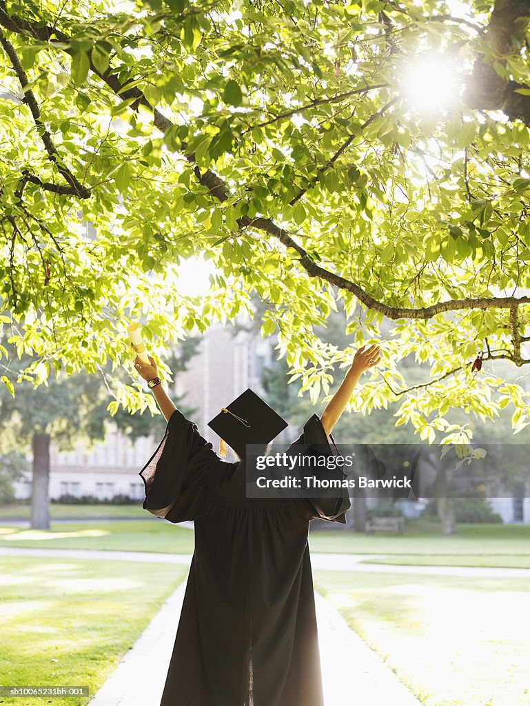 Young woman in graduation gown with raised arms outdoors, rear view