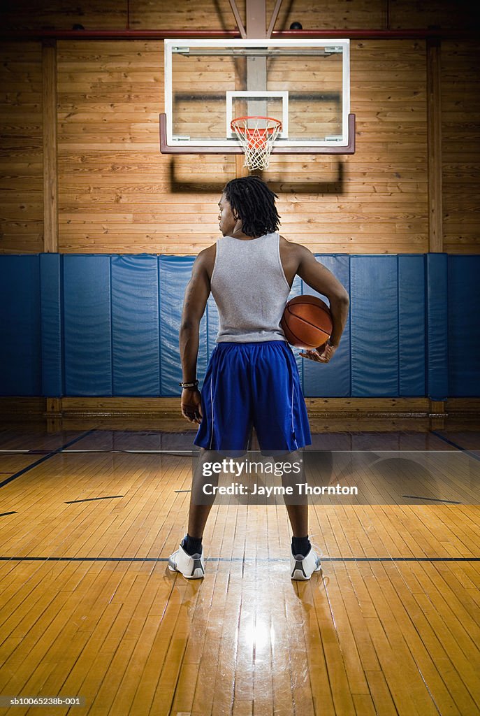 Man holding basketball under arm, standing on court, rear view