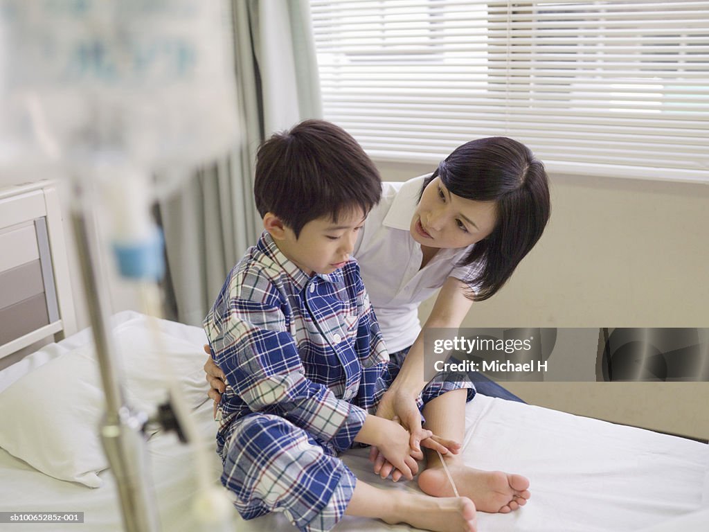 Boy (5-6) with drip, holding hands with mother on hospital bed