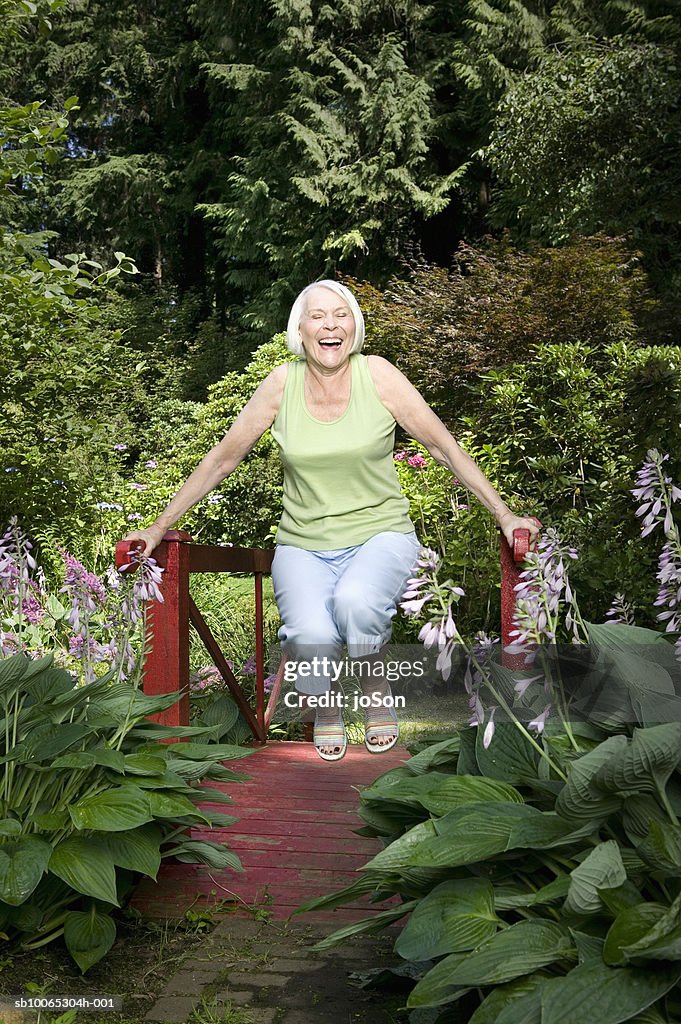 Senior woman swinging on small bridge in garden, laughing