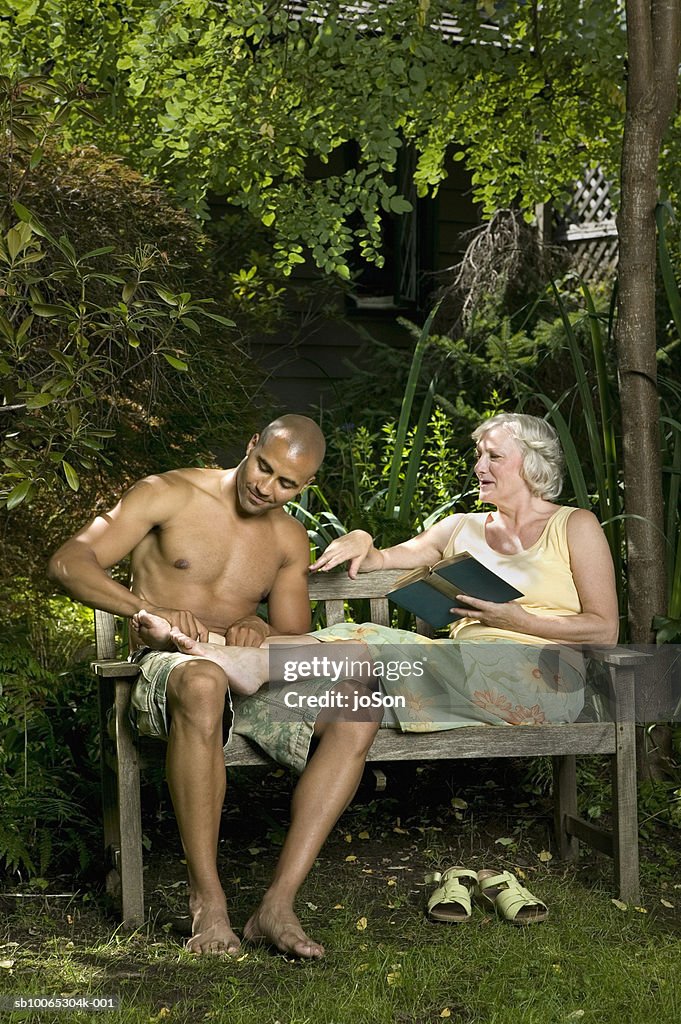 Young man and senior woman sitting on garden bench, man massaging woman's foot