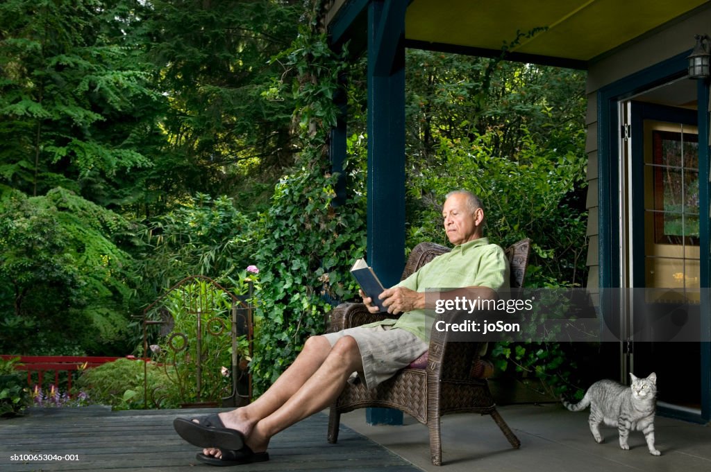 Senior man sitting on porch reading book, cat passing by