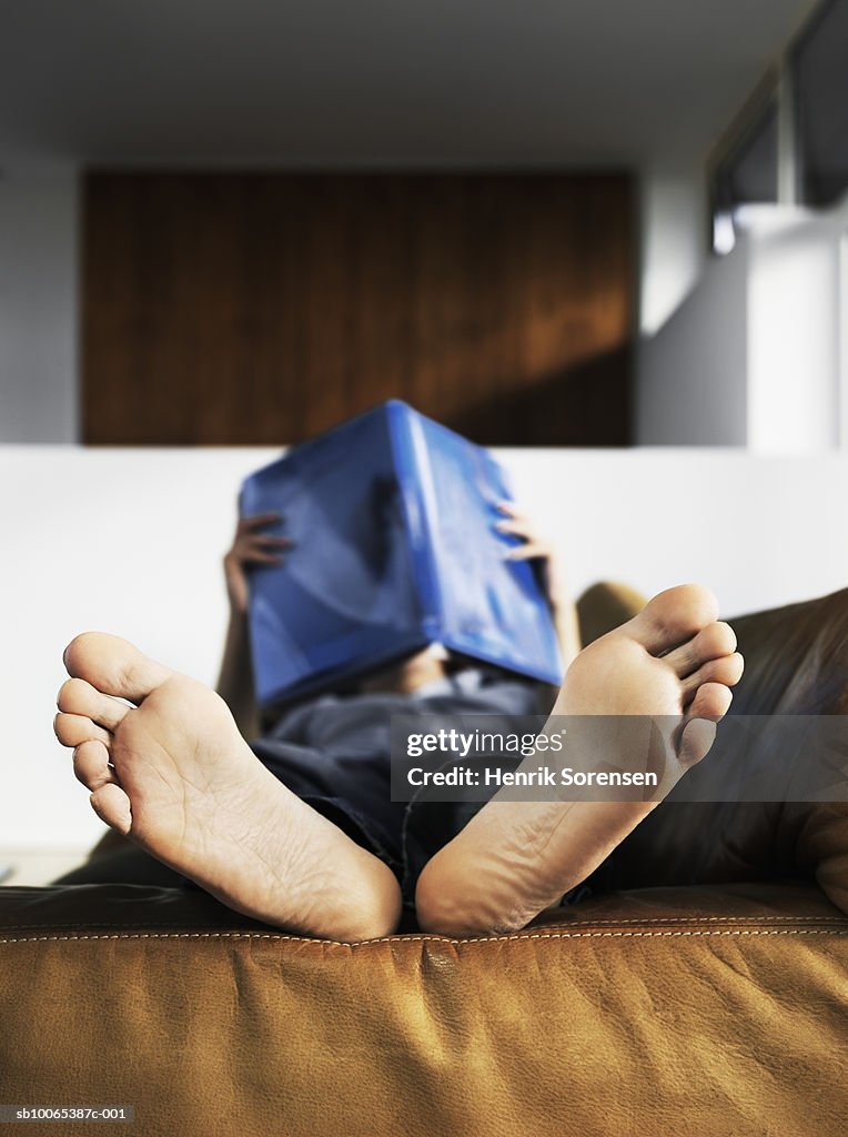 Mid adult man lying on sofa reading book, focus on barefoot